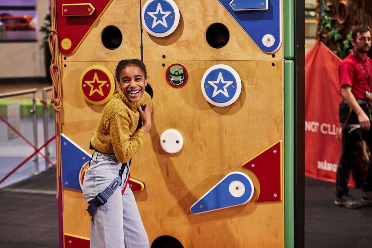 Child climber laughs, gripping a belay rope; colorful wall with star-decorated holds in an indoor climbing gym; an instructor stands near an orange safety banner.
Text: “NOT CLI...” (partial). Other small stickers have unreadable text.